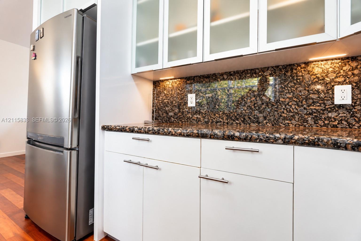 Kitchen area featuring white cabinetry with glass-front uppers, stone countertops, stone backsplash, stainless steel refrigerator, and wood-finish flooring