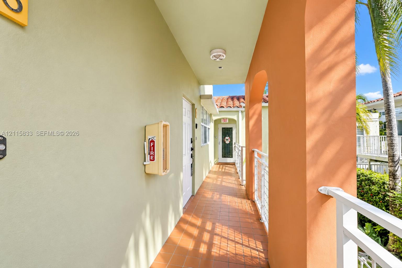 Covered exterior walkway featuring terra cotta tile flooring, stucco wall finishes, and structural archways with metal railings