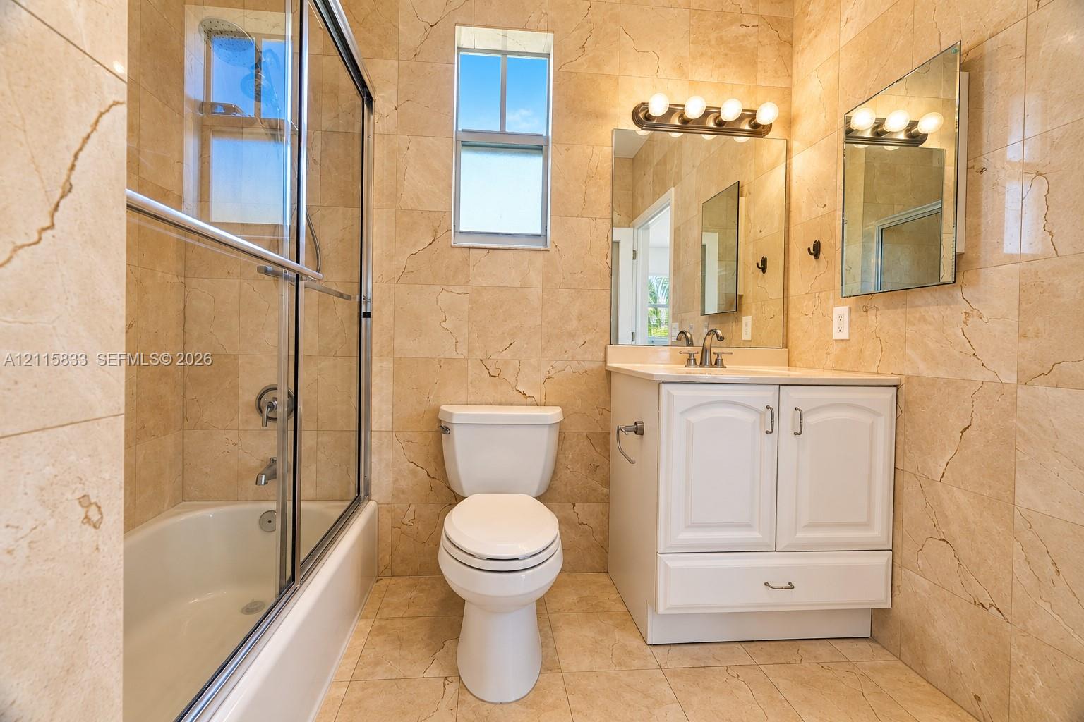 Floor-to-ceiling stone tile surrounds, white cabinetry with integrated hardware, glass-enclosed shower, wall-mounted multi-bulb light fixture, and large rectangular vanity mirror