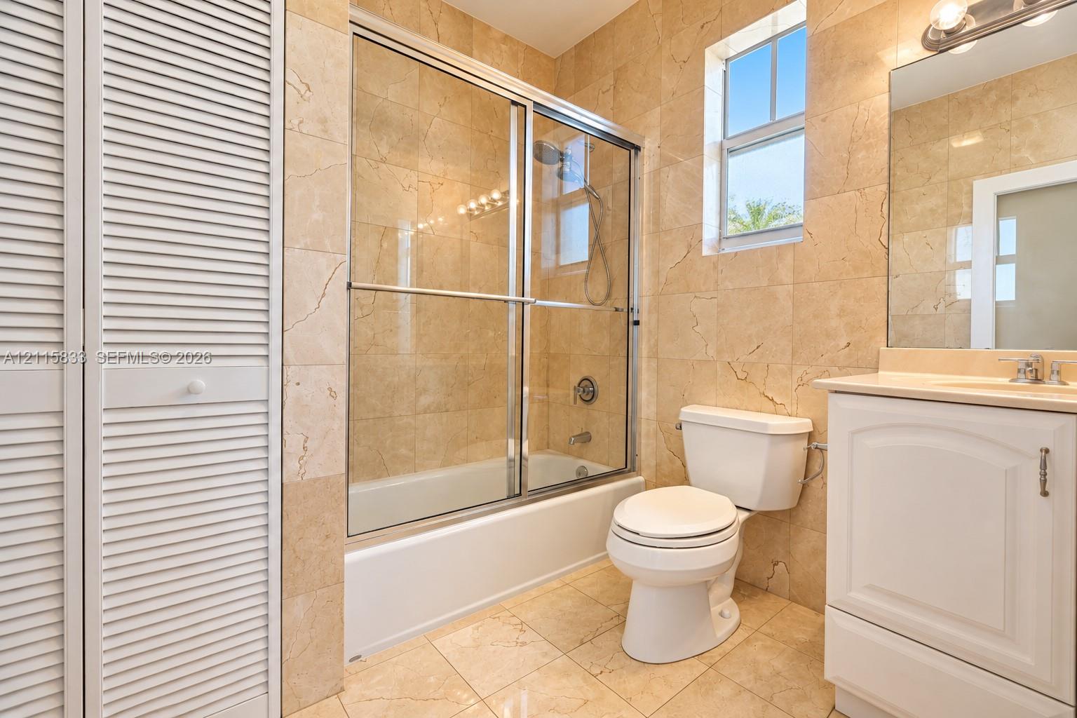 Bathroom featuring natural stone tile flooring and wall surfacing, a glass-enclosed shower and tub combination, a white vanity cabinet with a neutral countertop, and louvered closet doors
