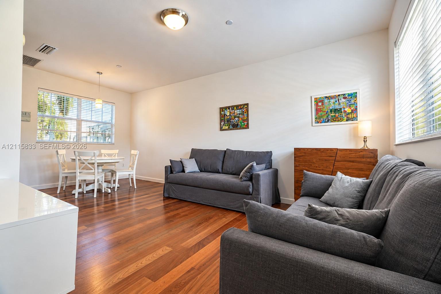 Open interior space featuring wood-finish flooring, dual windows with horizontal blinds, a suspended pendant light fixture, and a central flush-mount ceiling light