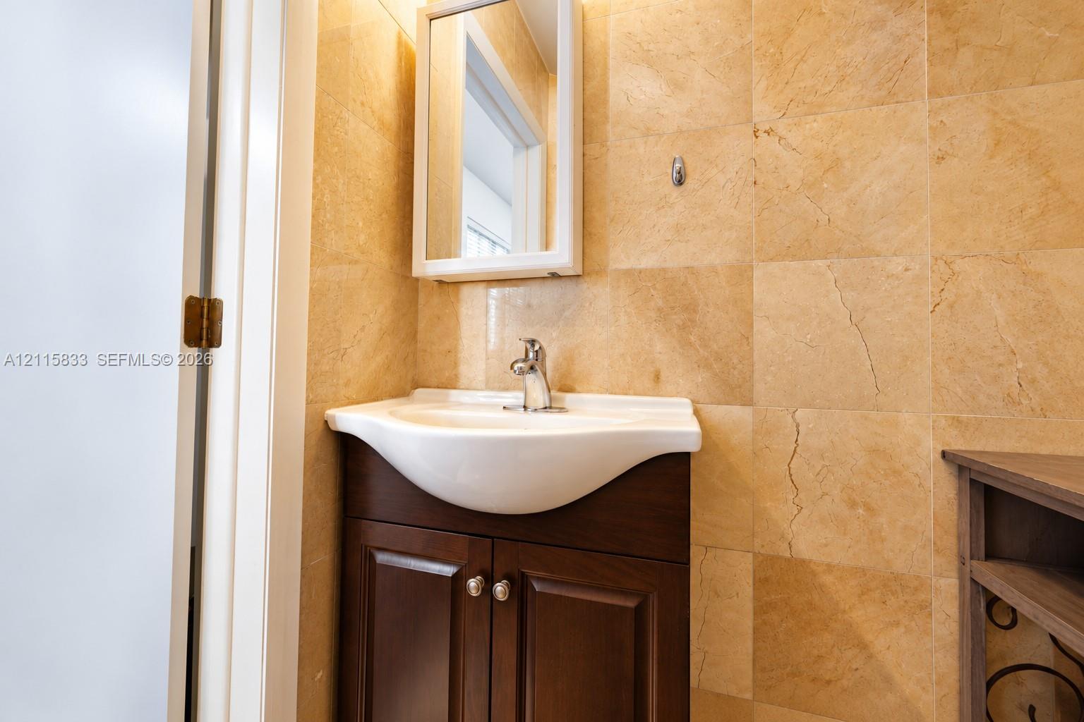 Wall-mounted medicine cabinet with a mirrored door, white pedestal-style sink basin, dark wood-finish cabinet, and full-height stone-finish wall tiling