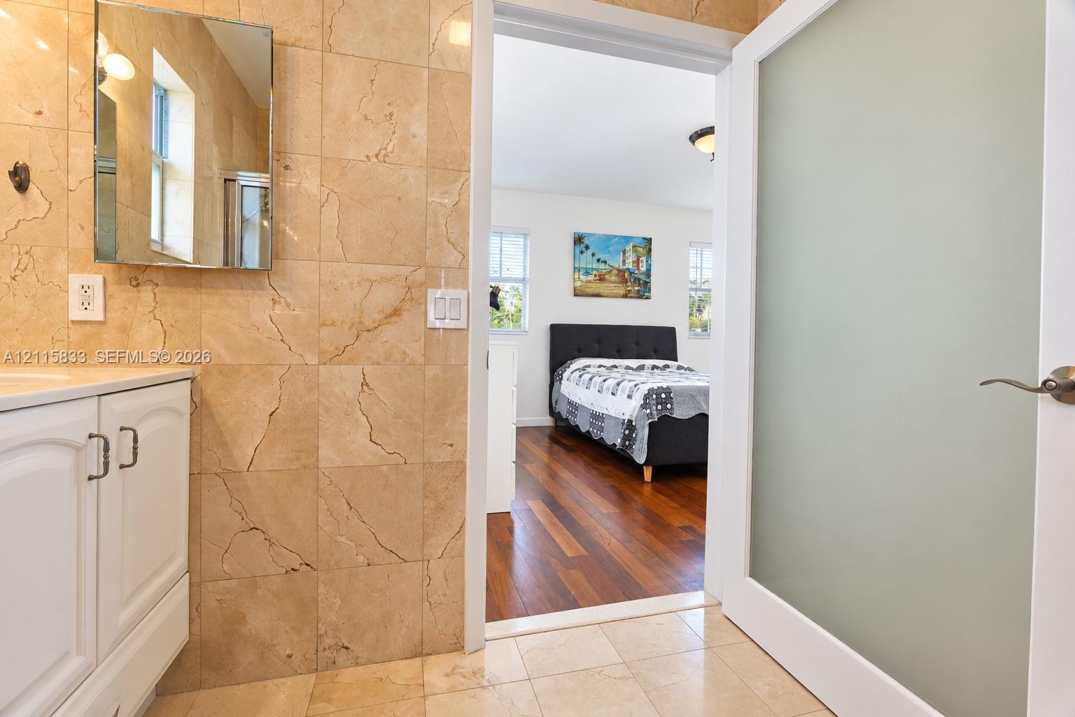Bathroom featuring large-format natural stone wall tiling, a white vanity with built-in cabinetry, and a frosted glass panel door with satin nickel hardware, opening to a space with wood-finish flooring