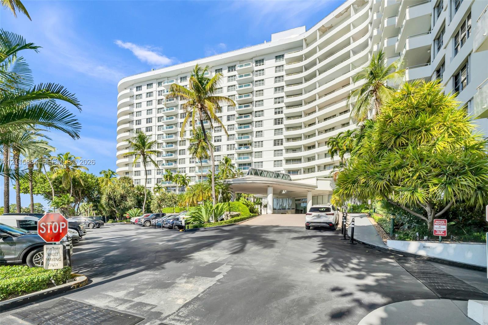Mid-rise exterior featuring curved structural architecture, stacked private balconies, a covered porte-cochre entry, and mature perimeter landscaping
