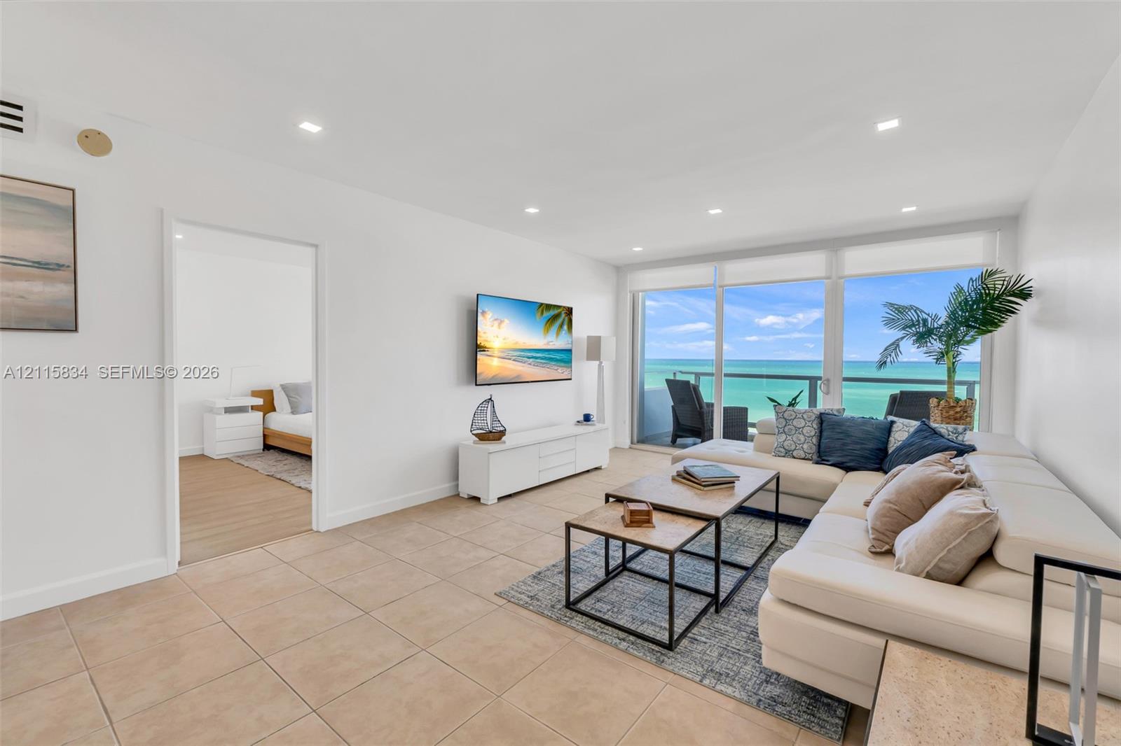 Living area featuring large-format neutral floor tiling, recessed lighting, and floor-to-ceiling glass doors opening to a balcony with an ocean backdrop
