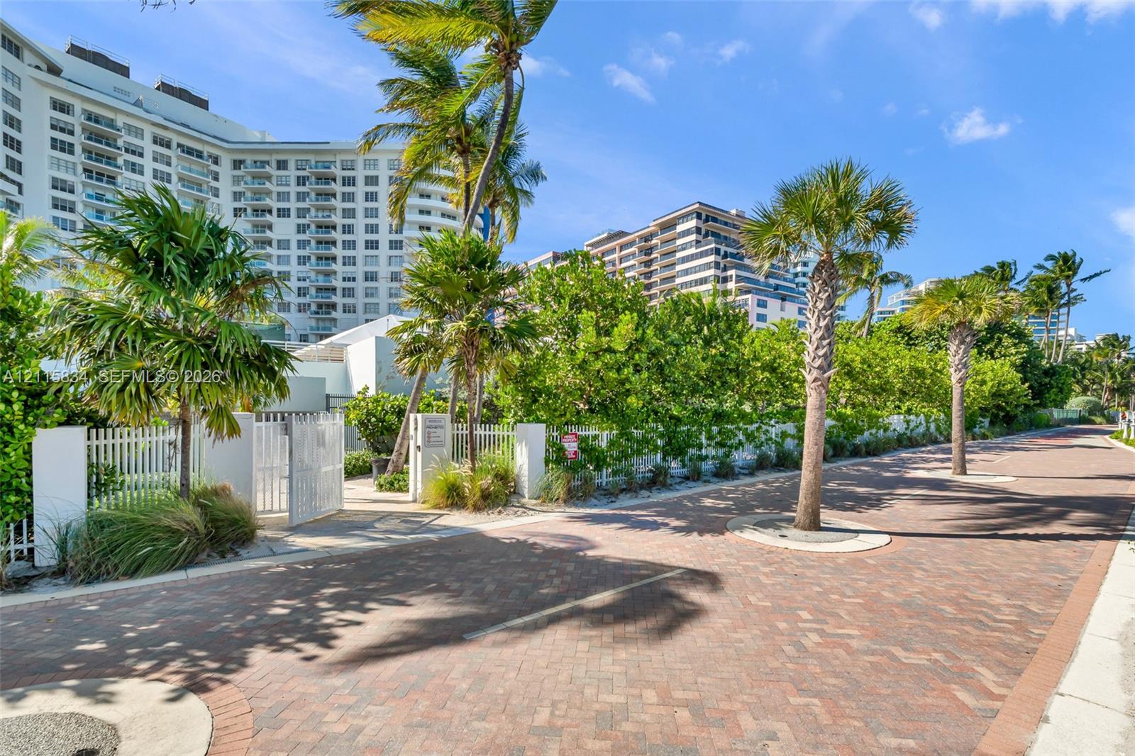 Brick-paved thoroughfare featuring mature palm trees, white metal fencing, and stone perimeter pillars