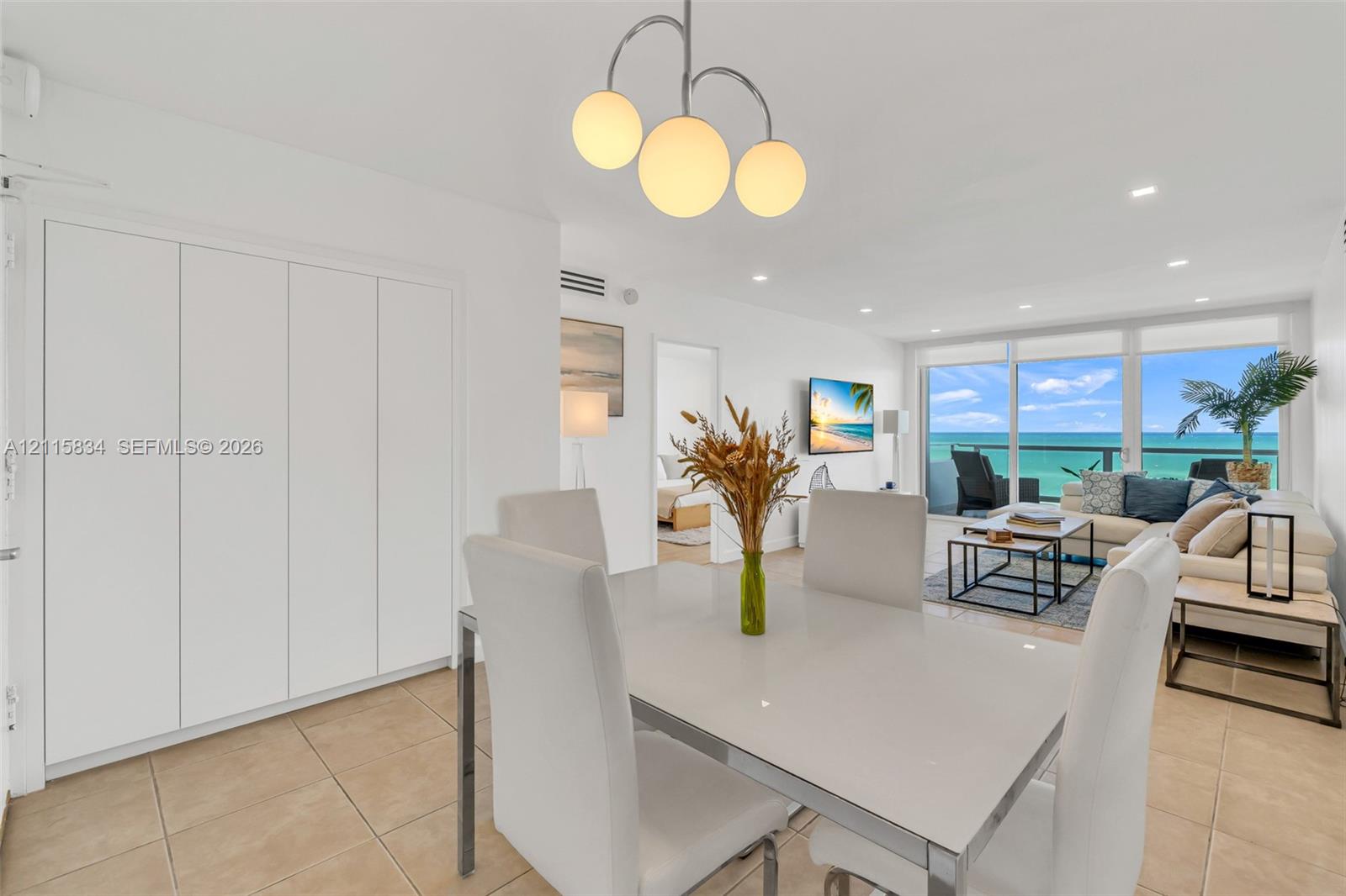 Open-plan living area featuring large-format tile flooring, built-in floor-to-ceiling cabinetry, recessed lighting, a modern three-globe pendant fixture, and glass sliding doors providing water access