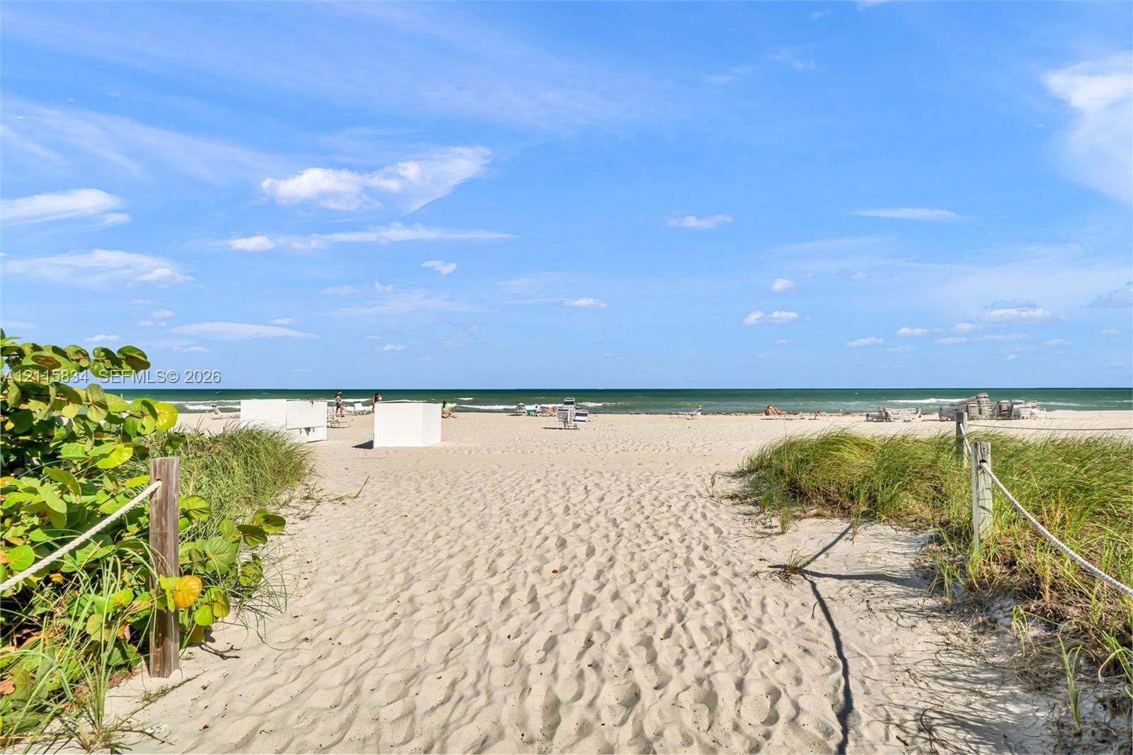 Sandy beach access featuring a sand-covered path, native coastal vegetation, and rope-style perimeter fencing
