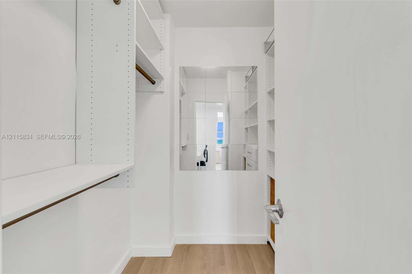 Walk-in closet featuring white built-in shelving, integrated hanging rods, wall-mounted mirror, and wood-finish flooring