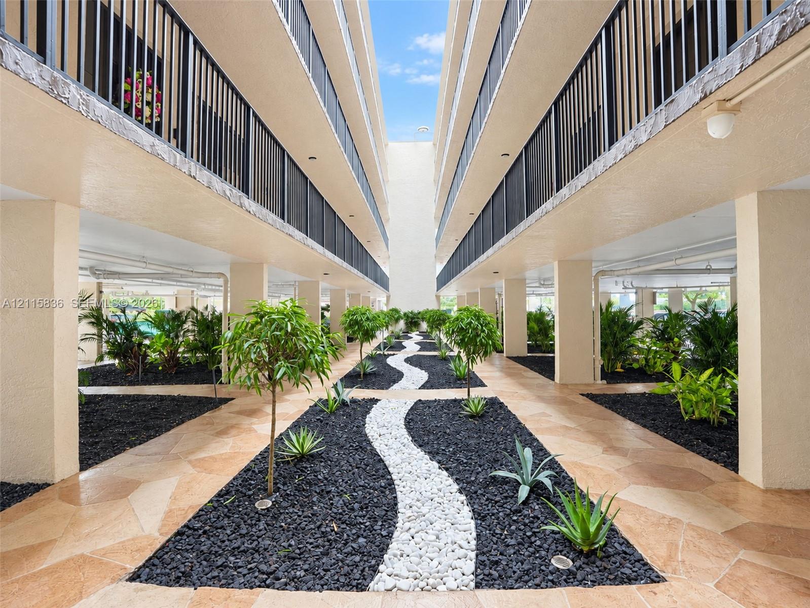 Multi-story building featuring structural columns, decorative black metal railings, stone-paver ground surfaces, and manicured landscaping with organic rock borders