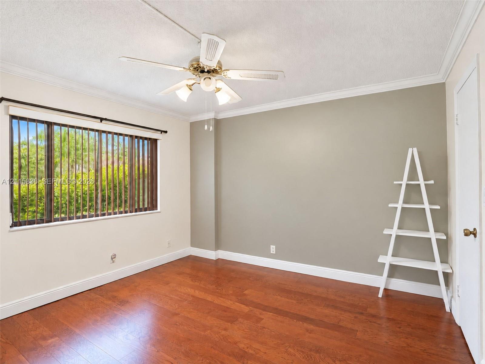 Room featuring wood-finish flooring, a multi-blade ceiling fan with integrated lighting, crown molding, and a wide window with vertical blinds