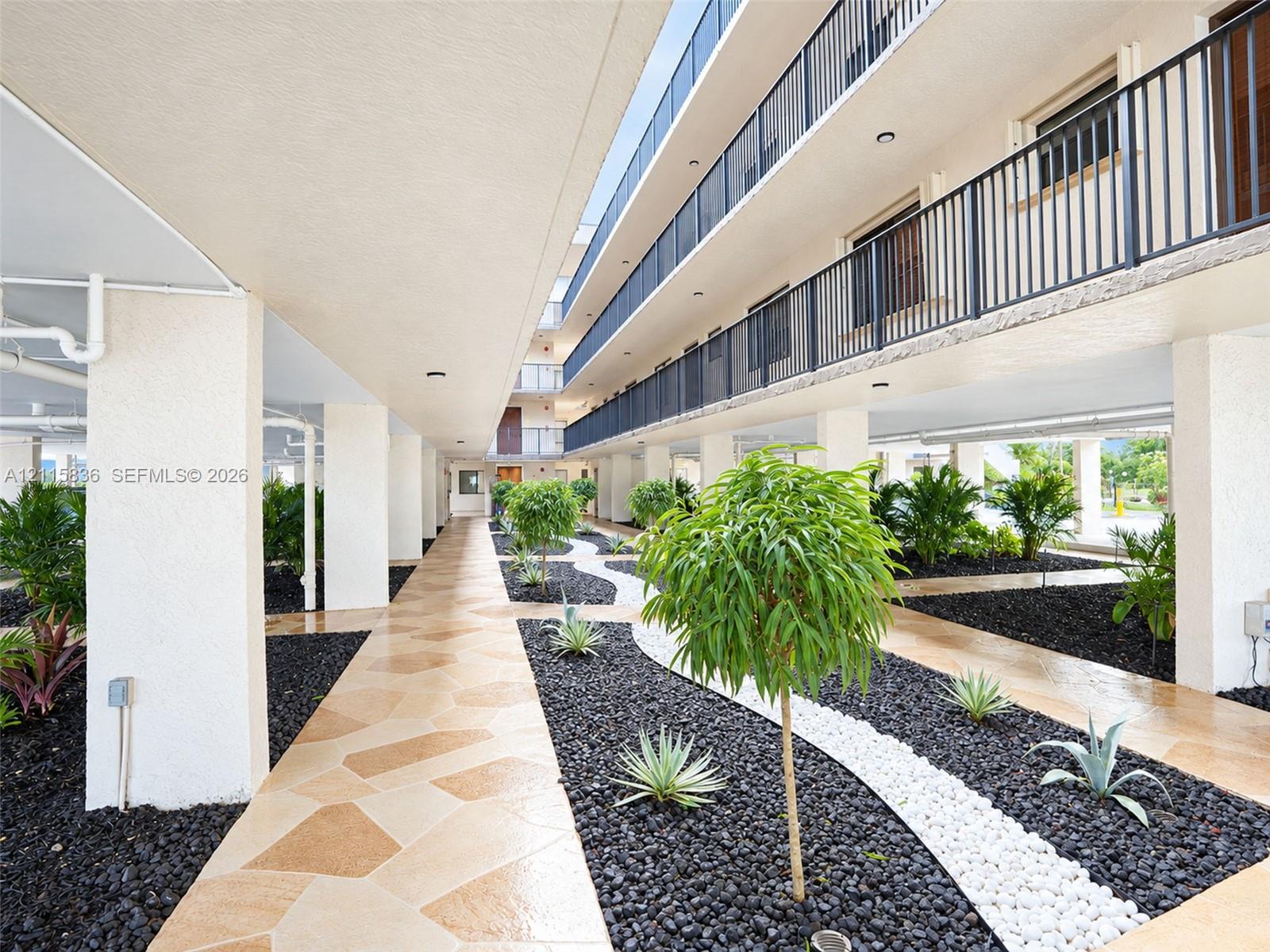 Covered ground-level walkway featuring paver stone pathways, dark volcanic rock landscaping, white decorative stone borders, integrated ornamental shrubbery, and structural concrete pillars
