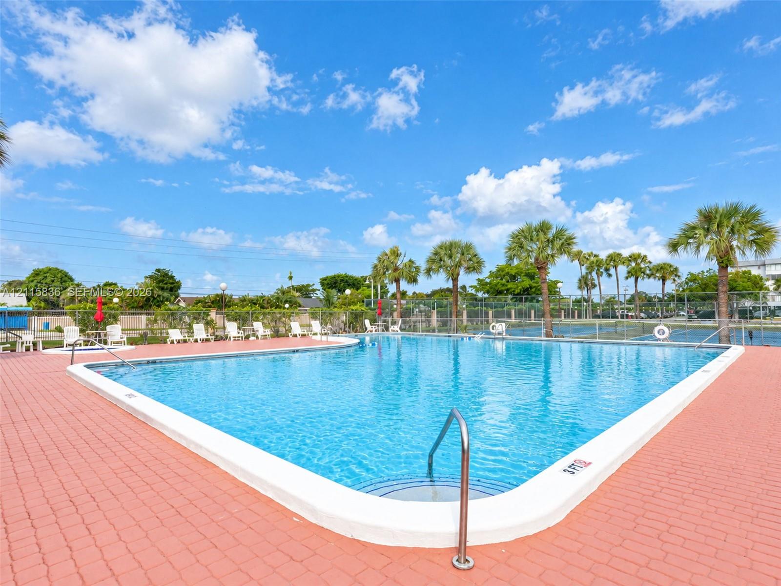 Outdoor swimming pool featuring a red brick paver deck, metal entry railings, and surrounding palm trees