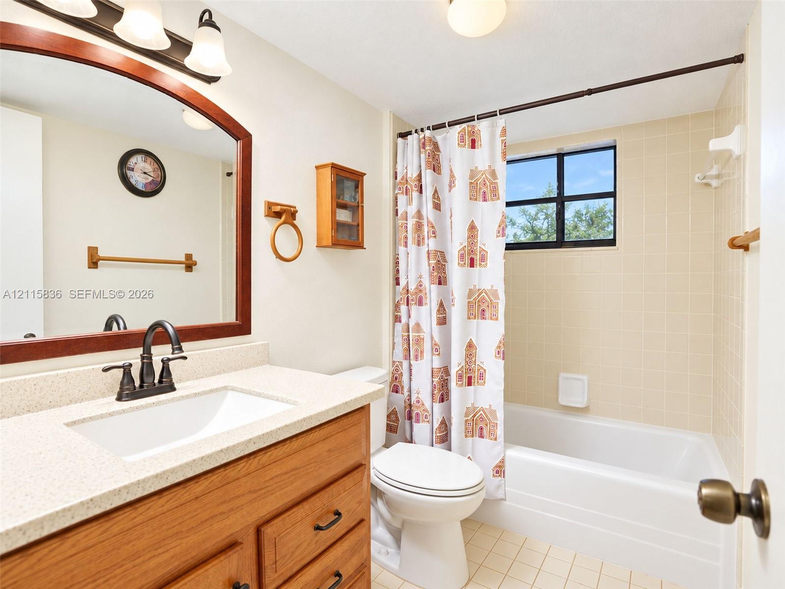 Bathroom featuring a wood-tone vanity, light-colored stone-finish countertop with an undermount sink, oil-rubbed bronze fixtures, arched wall-mounted mirror, and a tiled tub-shower combination with a window