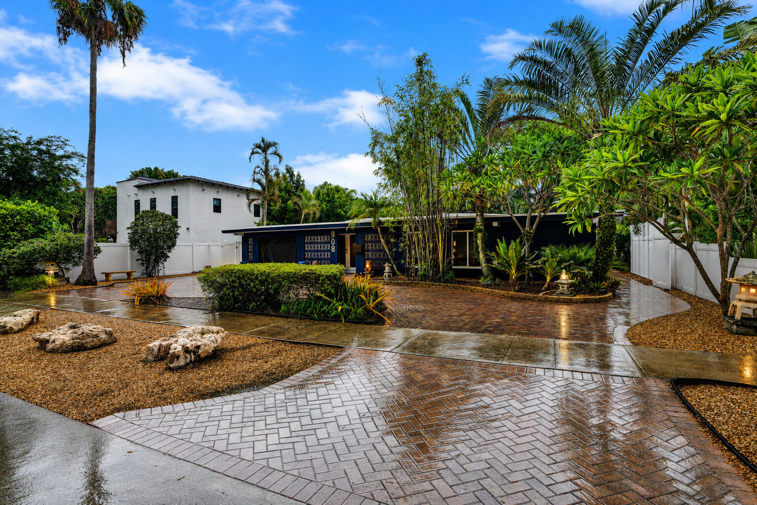 Paved driveway featuring a herringbone pattern, gravel landscaping with natural rock elements, mature palm trees, and lush tropical foliage Paved driveway featuring a herringbone pattern, gravel landscaping with natural rock elements, mature palm trees, and lush tropical foliage