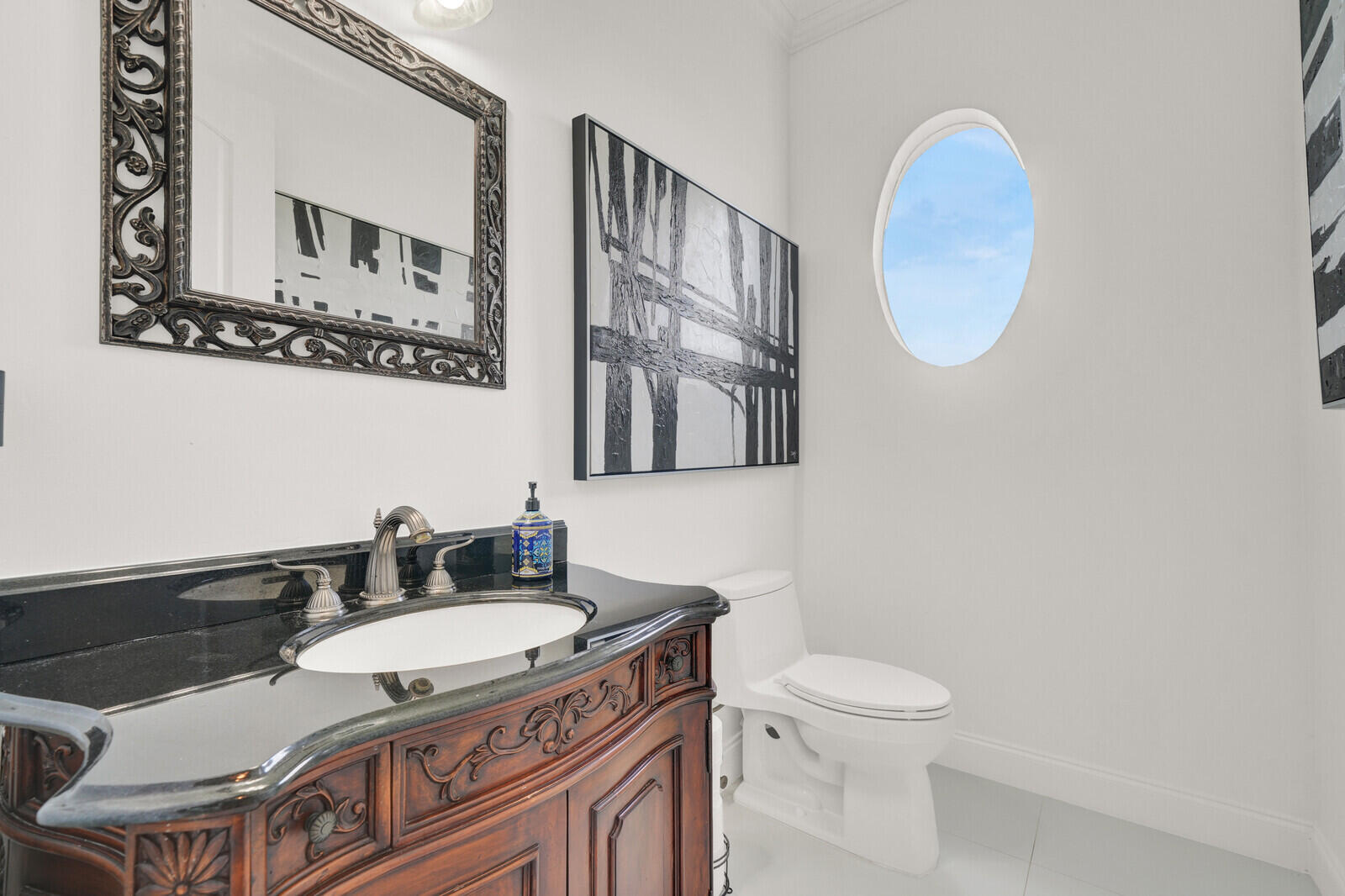 Powder room featuring an ornate wood vanity with a black countertop, a decorative framed mirror, and an oval window