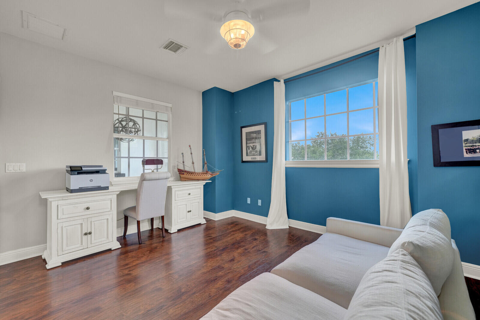 Hardwood-finish flooring anchors a room featuring a large window with white trim, a built-in white desk with cabinets, and a ceiling fan with integrated lighting