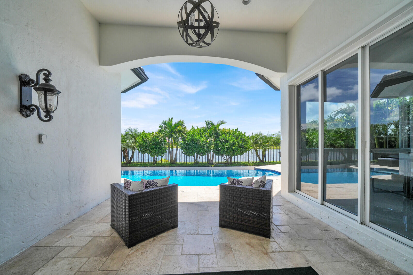 Covered patio with stone tile flooring, an arched entryway, and an ornate ceiling light fixture, opening to a private swimming pool and natural green landscaping