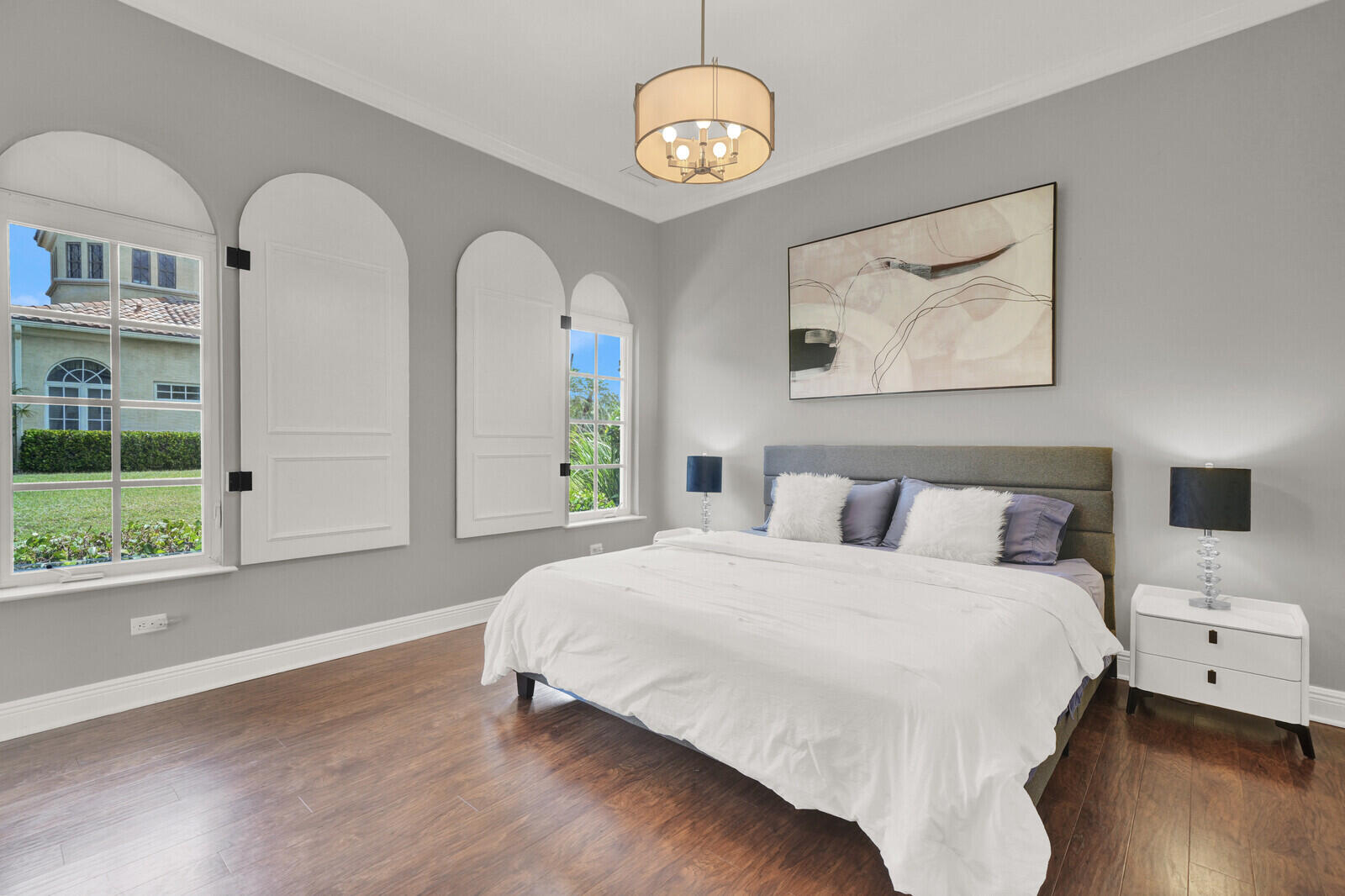 Expansive bedroom featuring arch-top windows with decorative interior shutters, rich wood-finish flooring, and a contemporary drum pendant light fixture