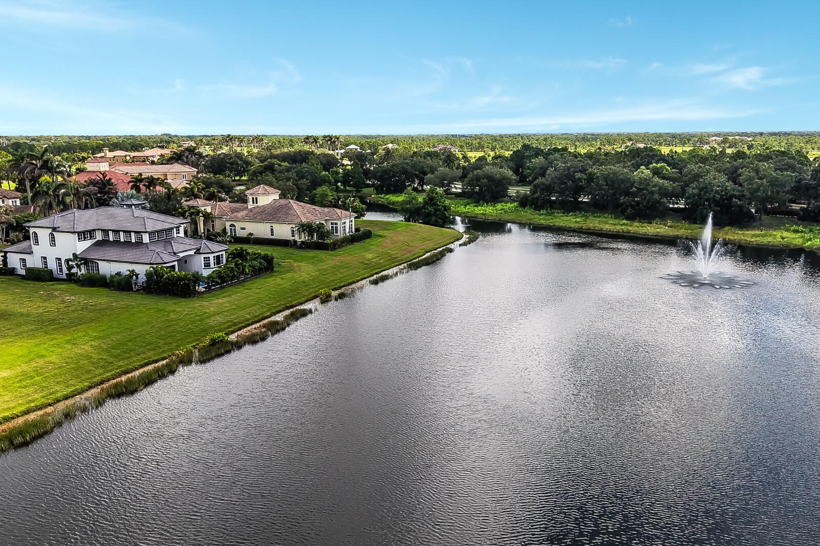 Expansive waterfront property featuring a meticulously landscaped lawn, mature trees, and a tranquil water body with a central fountain