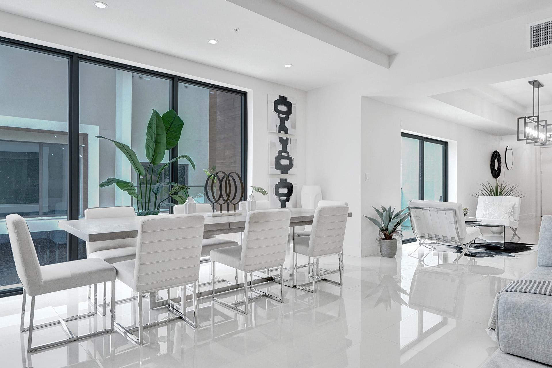 Sophisticated open-concept dining and living area featuring polished light-toned flooring, floor-to-ceiling windows with dark-framed glazing, contemporary white upholstered dining chairs with metal frames, and a sleek white dining table