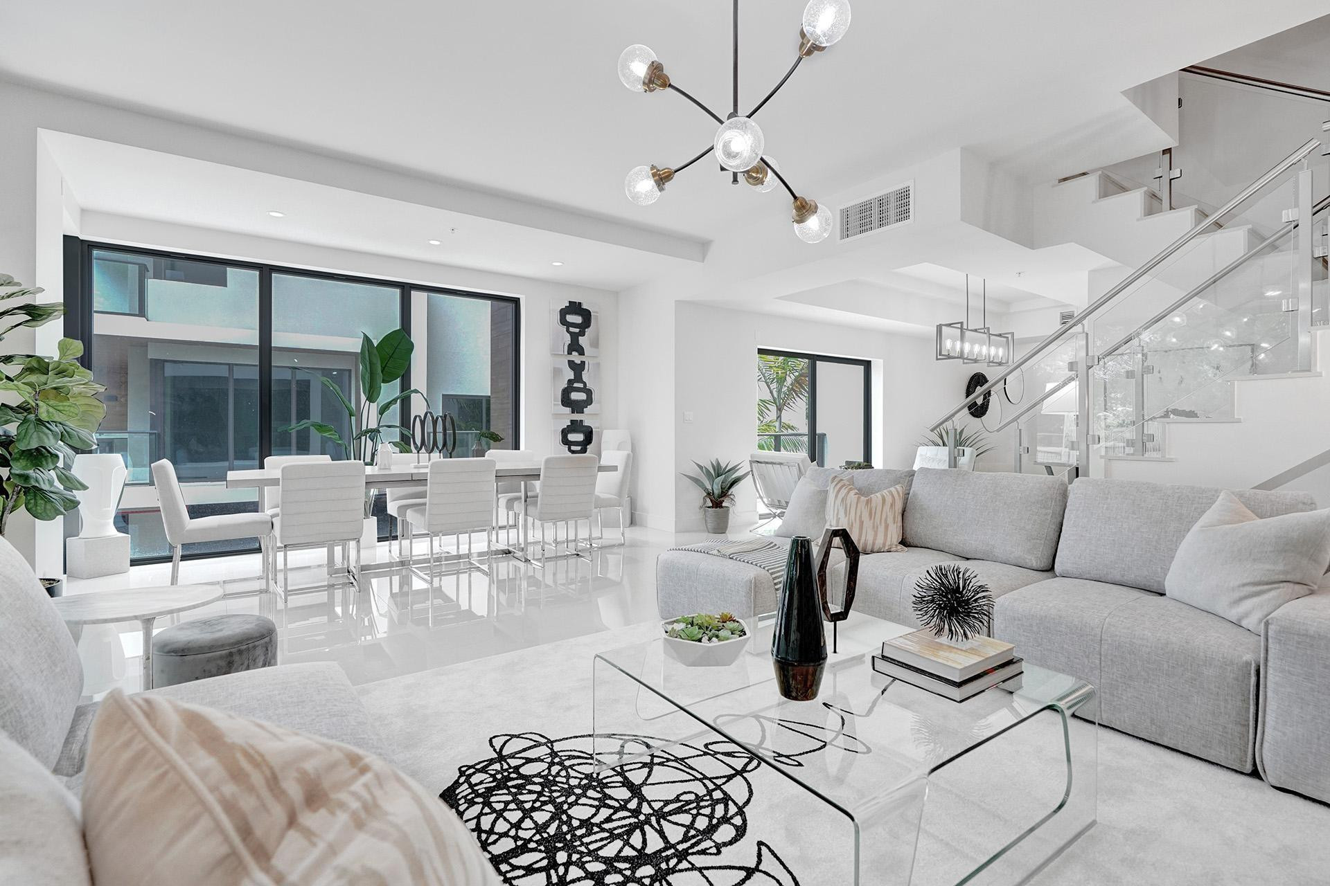 Spacious open-concept living area featuring a modern chandelier, polished tile flooring, and a glass-paneled staircase with a metal handrail