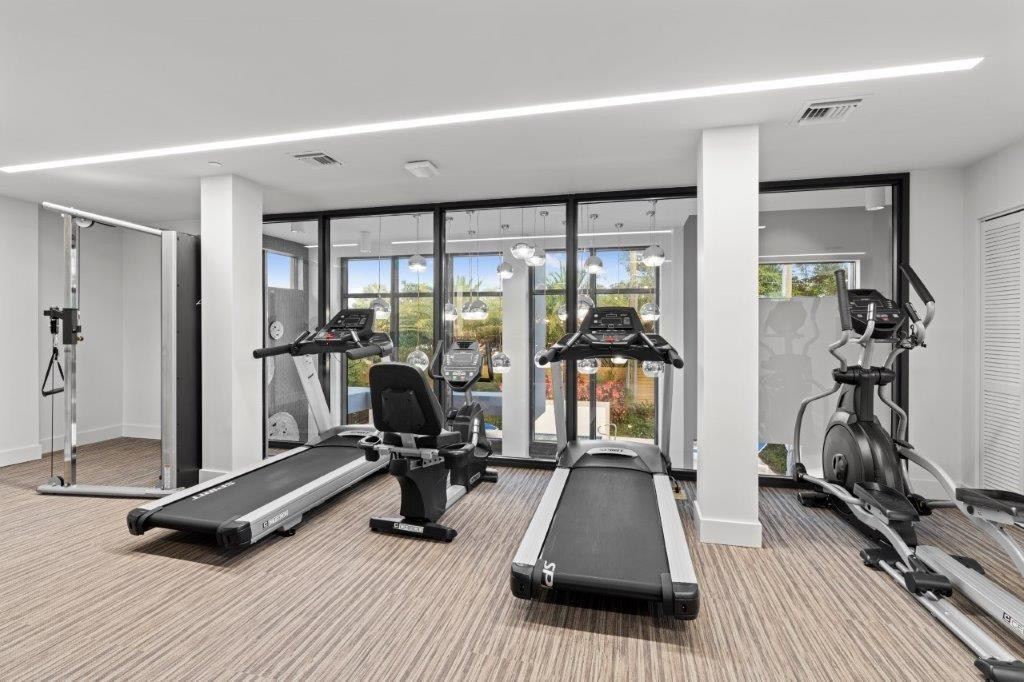 State-of-the-art fitness center featuring multiple cardio and strength-training stations, including treadmills and stationary bikes, anchored by support columns and bathed in natural light from expansive glass doors and windows