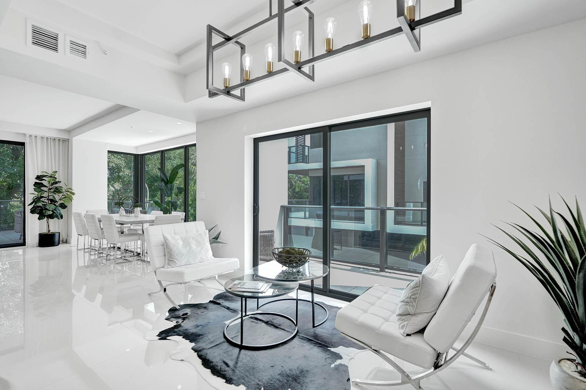 Spacious living area featuring polished white flooring, a modern linear chandelier with exposed bulbs, and a sliding glass door opening to a private balcony