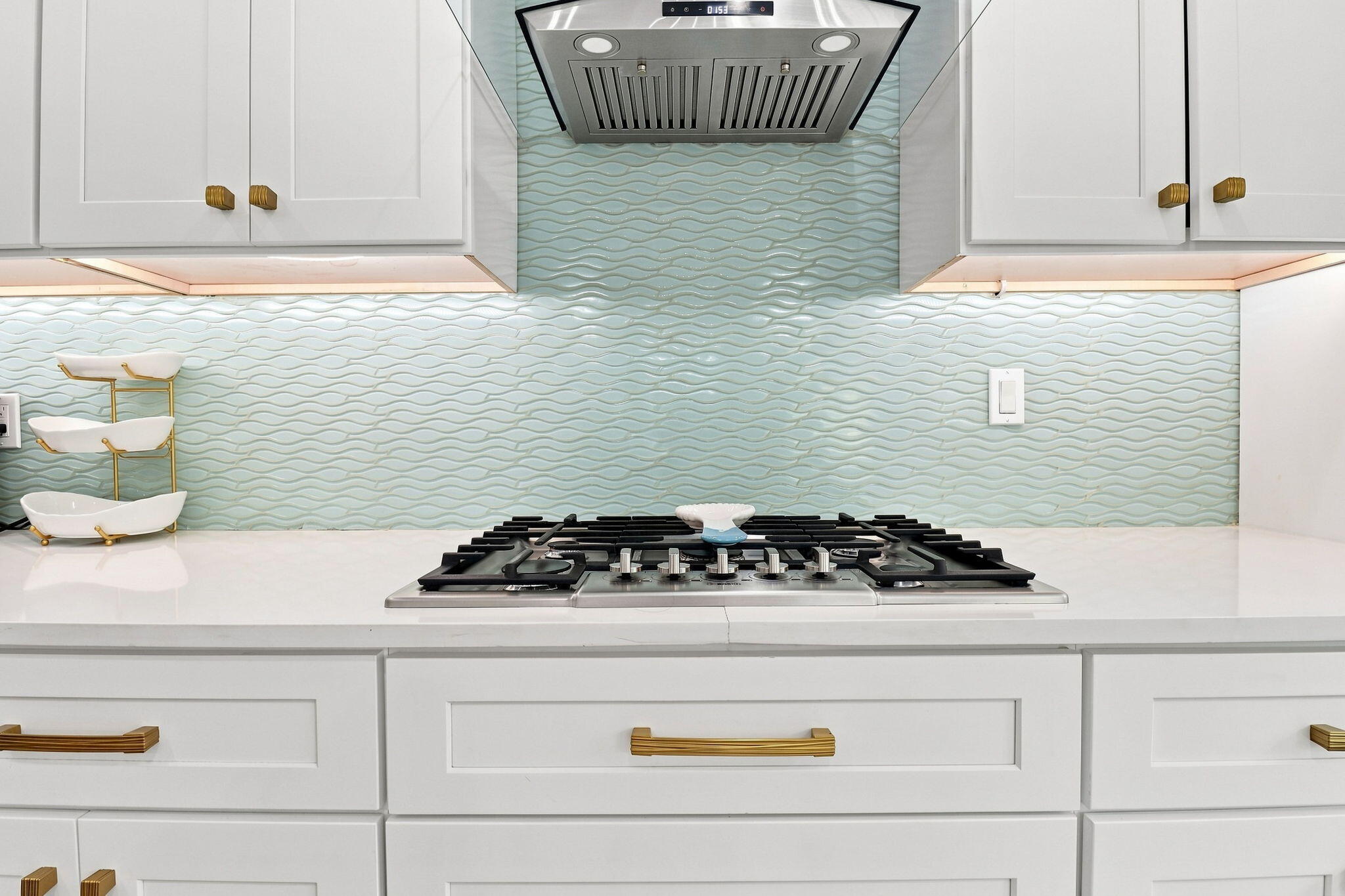 White shaker cabinetry with gold-tone hardware, built-in cooktop, stainless steel range hood, light-toned countertop and a wave-patterned tile backsplash