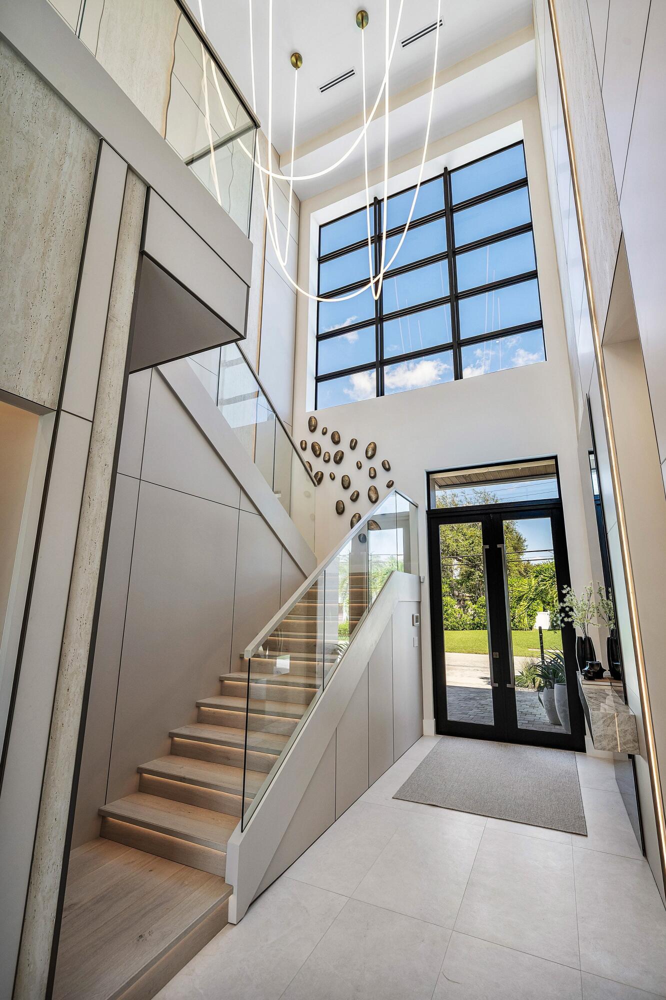 Glass-paneled double entry doors, high ceiling, upper-level windows, modern staircase with glass railing, and integrated lighting