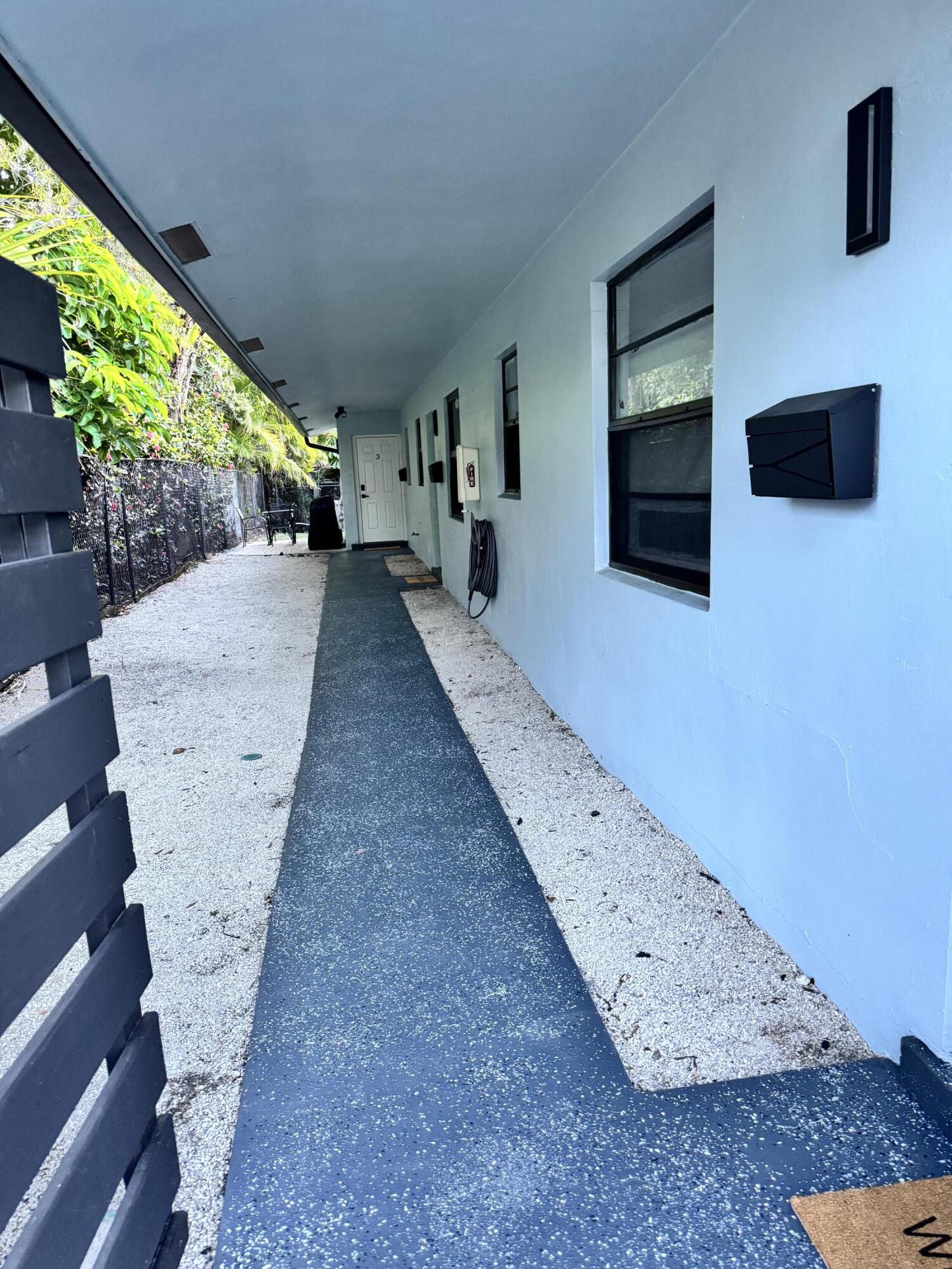 Covered walkway with a blue-painted concrete path, white stucco exterior, black-framed windows, and a modern black mailbox Covered walkway with a blue-painted concrete path, white stucco exterior, black-framed windows, and a modern black mailbox
