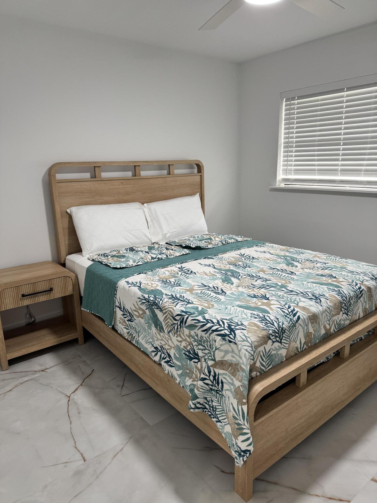 Bright and inviting bedroom featuring a modern wood bed frame, coordinating nightstand, tile flooring, and a window with blinds.
