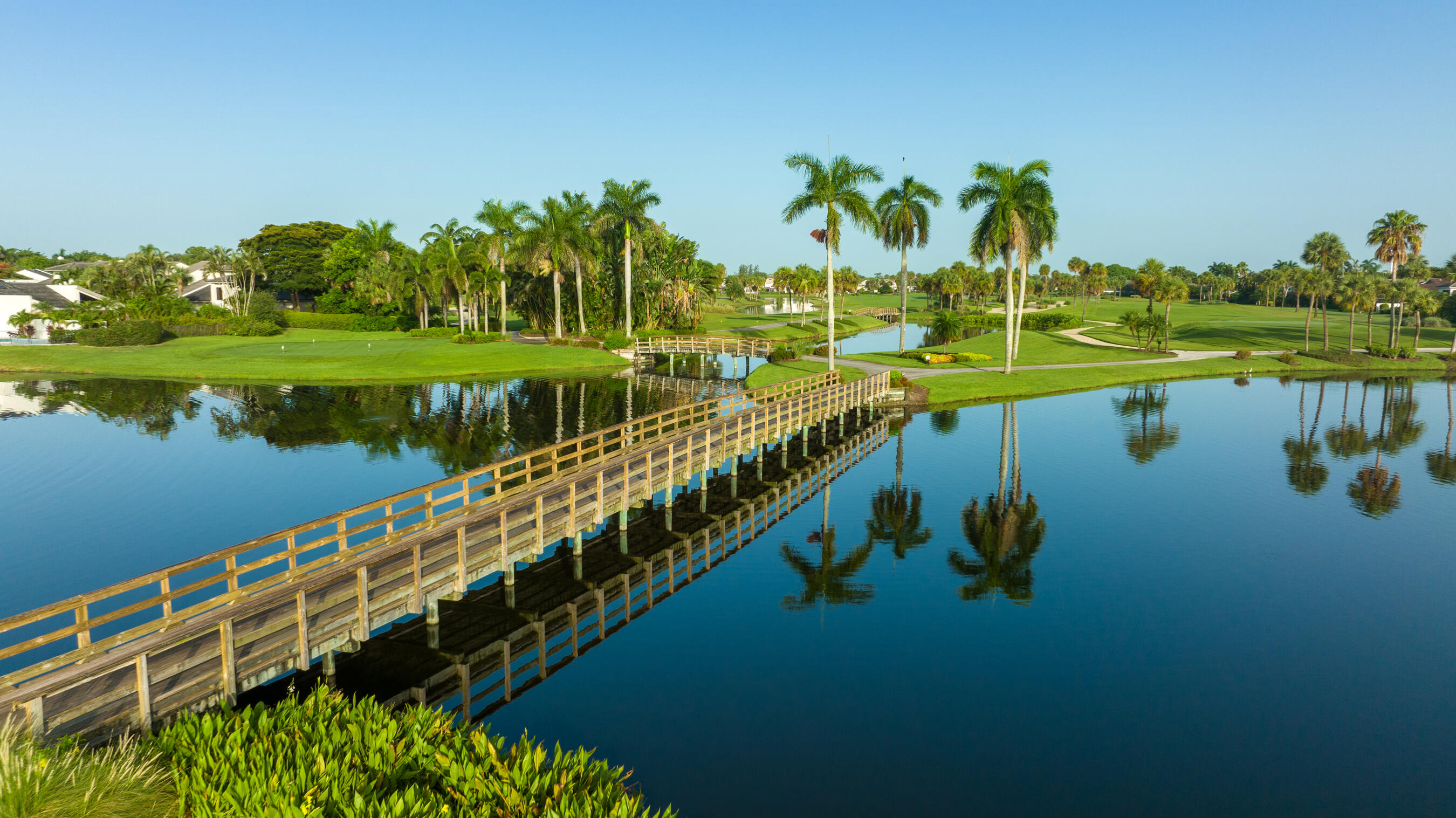 Serene waterfront setting featuring a wooden boardwalk spanning tranquil waters, lush landscaping with mature palm trees, manicured lawn areas, and additional waterfront parcels visible across the property grounds