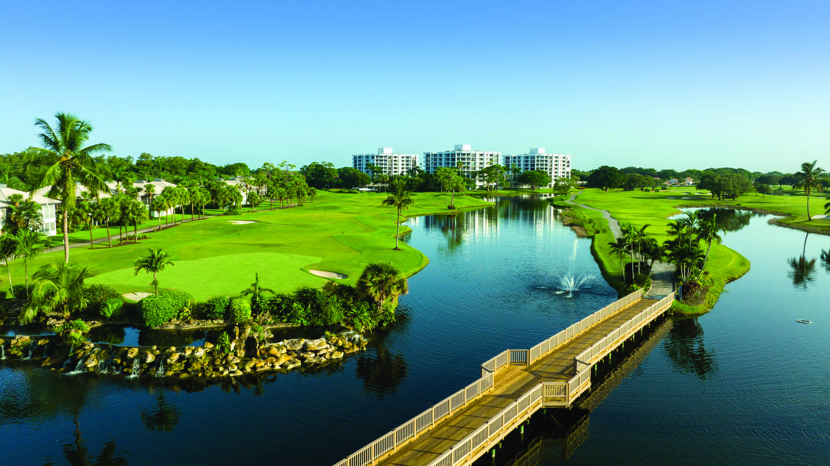 Expansive golf course with mature palm trees, multiple water features, a pedestrian bridge, and distant residential buildings