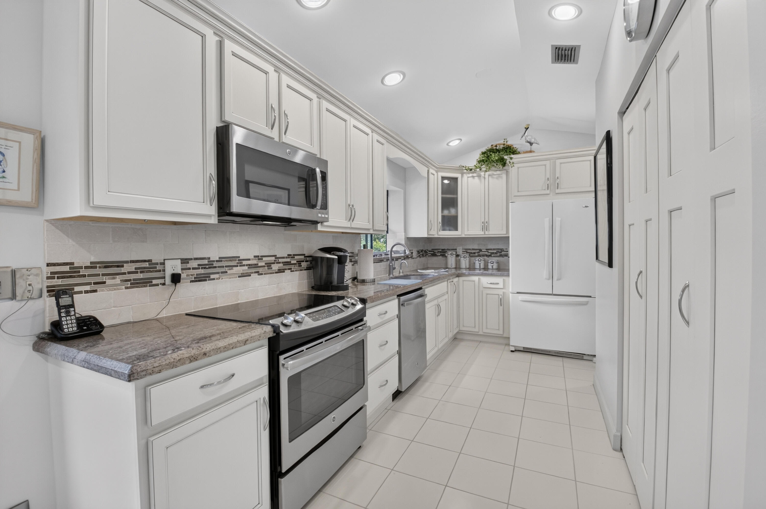 Vaulted ceiling kitchen featuring white cabinetry, tile flooring, stone countertops, stainless steel microwave, and integrated panel pantry storage