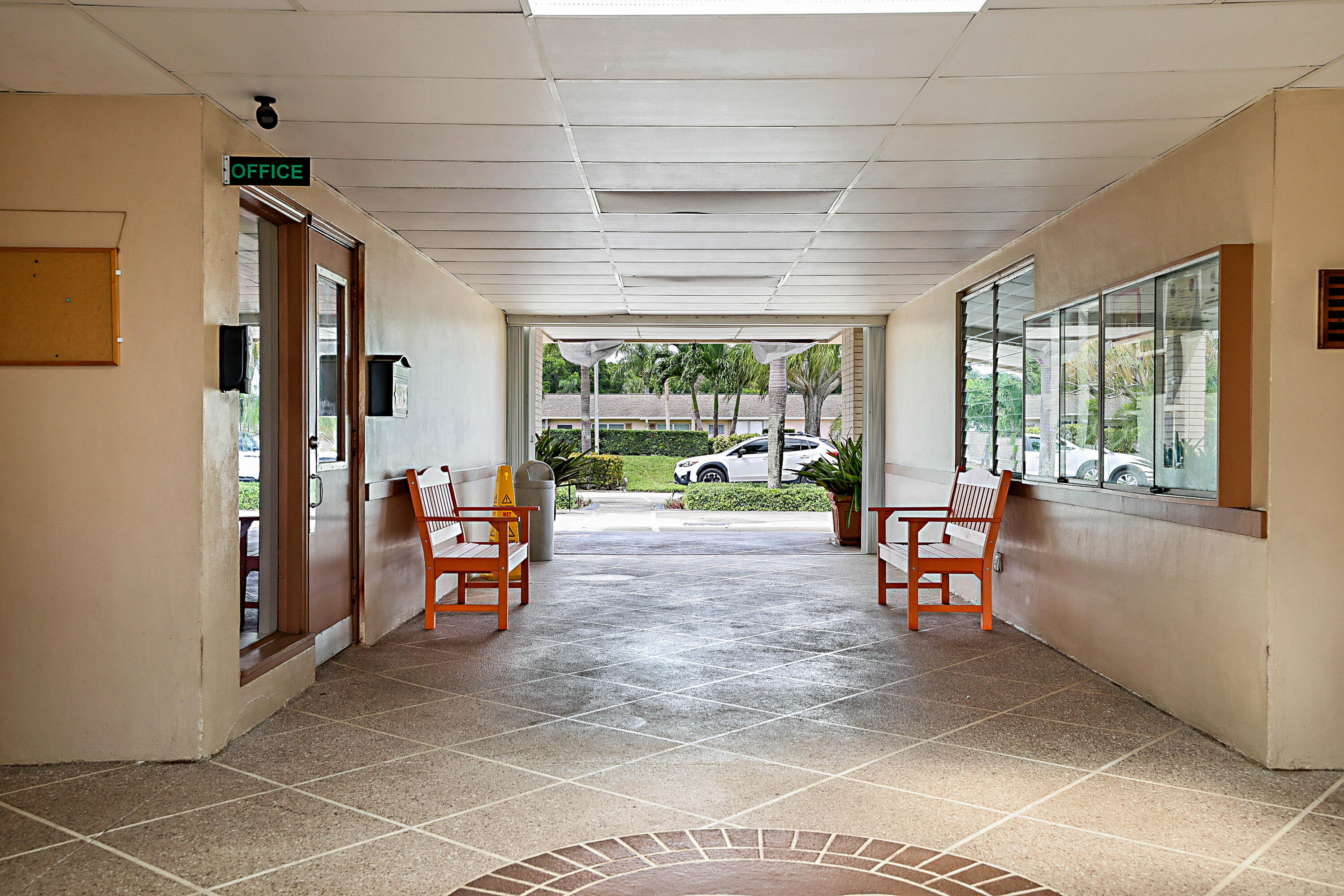 Covered walkway with a stone-finish tile floor featuring an inlaid brick pattern, providing access to managment office with a glass-paneled door