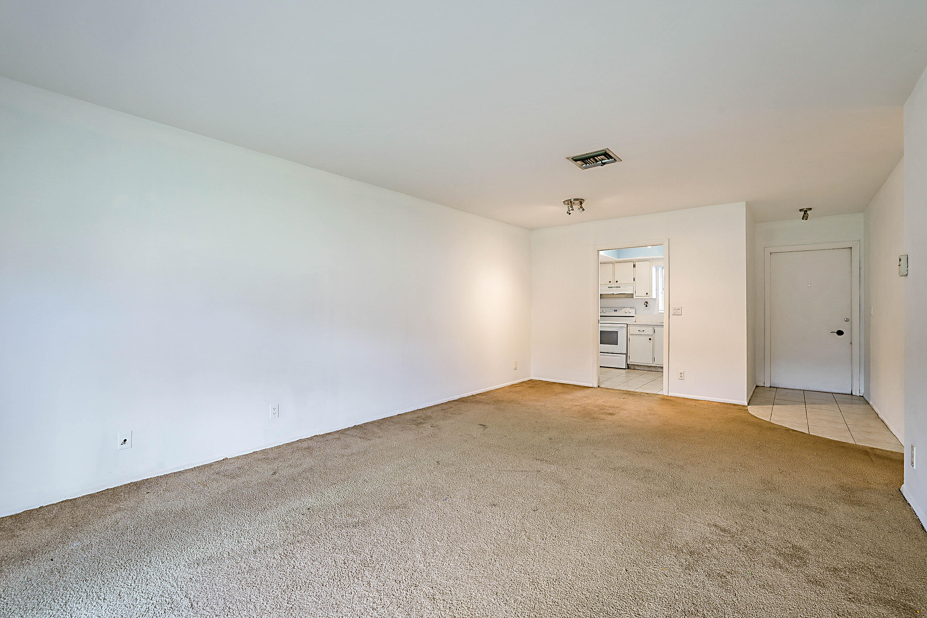 Carpeted living area with white walls and a doorway leading to a kitchen with white appliances