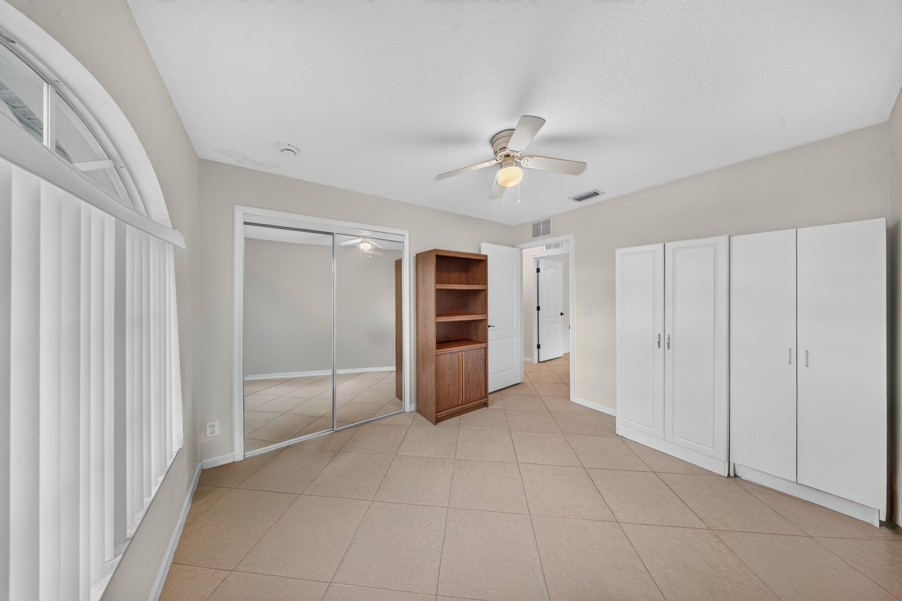 Tiled flooring, overhead ceiling fan, mirrored closet doors, built-in white cabinetry, and an arched window with vertical blinds