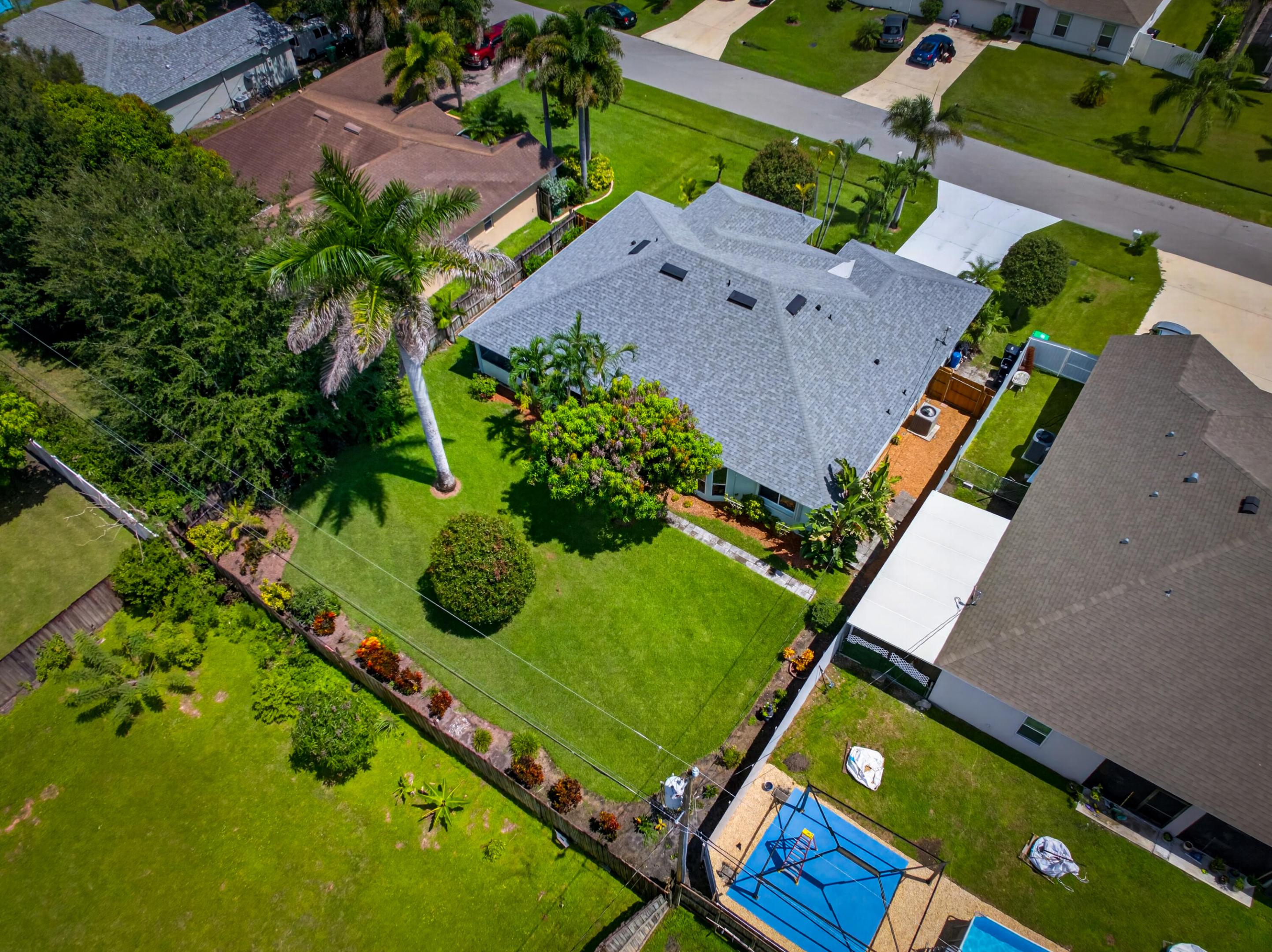 Single-story residence with a gray shingle roof, expansive green lawn, mature palm trees, landscaped garden beds, and a paved driveway