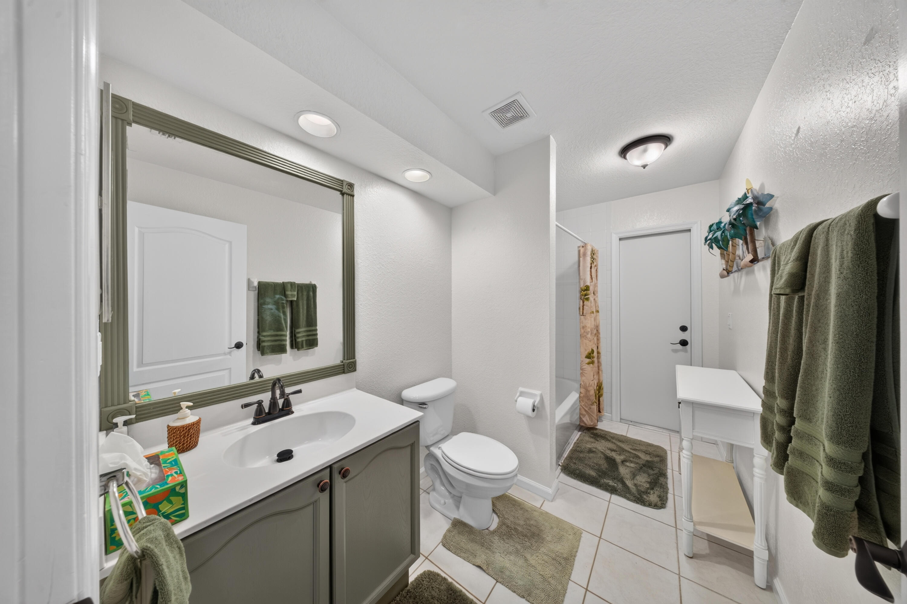 White tile flooring, single vanity with light countertop and gray cabinetry, framed mirror, recessed lighting, and a built-in bathtub with a shower