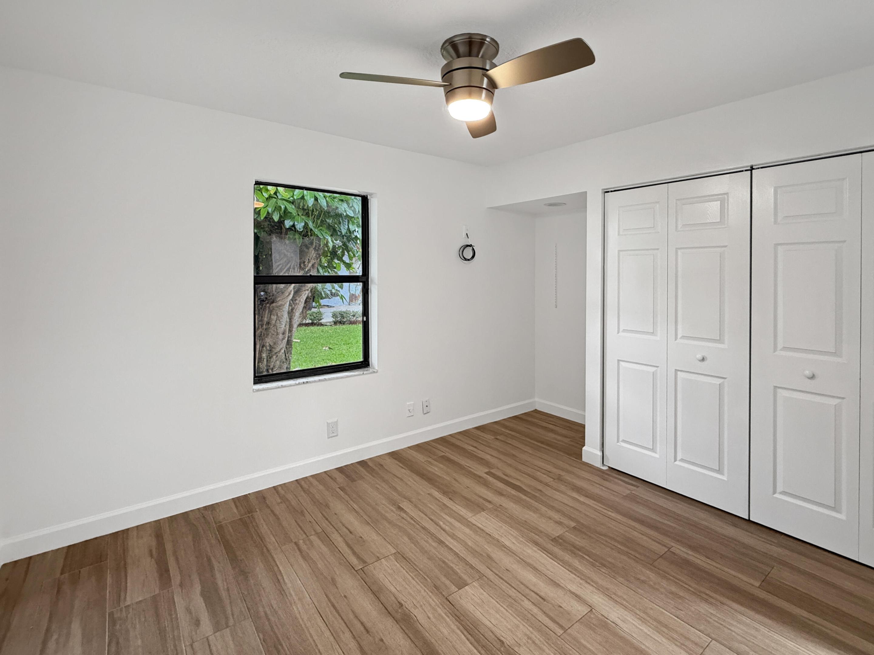 Spacious bedroom showcasing warm wood-finish flooring, white six-panel closet doors with multiple storage sections, ceiling-mounted fan with integrated lighting, and abundant natural light from a large window framing outdoor scenery