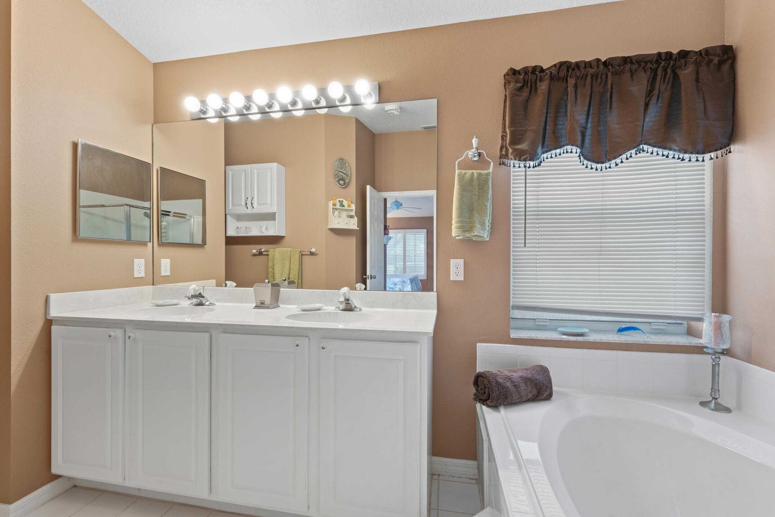 Dual-sink vanity with white cabinetry and solid surface countertop, large wall-mounted mirror, integrated strip lighting, and soaking tub with white tile surround