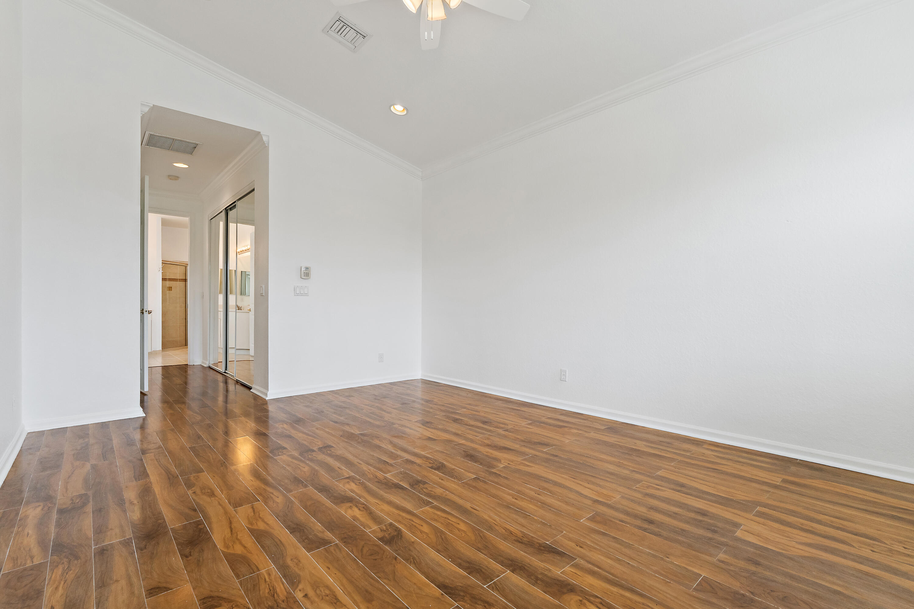 The primary bedroom has plenty of room for a king bed and sitting area, with wood-look floors, crown molding, and a ceiling fan. A short hallway leads to the walk-in closet and private bath, giving the suite a quiet, separate feel.