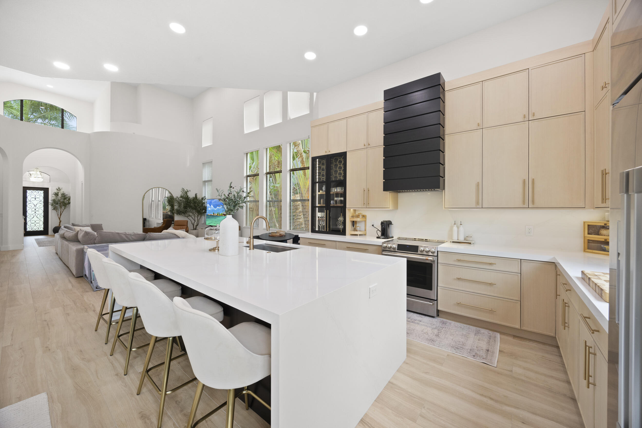 Gourmet kitchen featuring a large quartz-topped waterfall island, light wood cabinetry, a striking tiered black range hood, high ceilings, and wood-finish flooring
