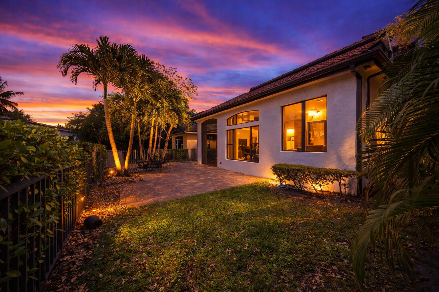 Stucco exterior featuring a paver patio, expansive windows, mature palm trees, and metal fencing