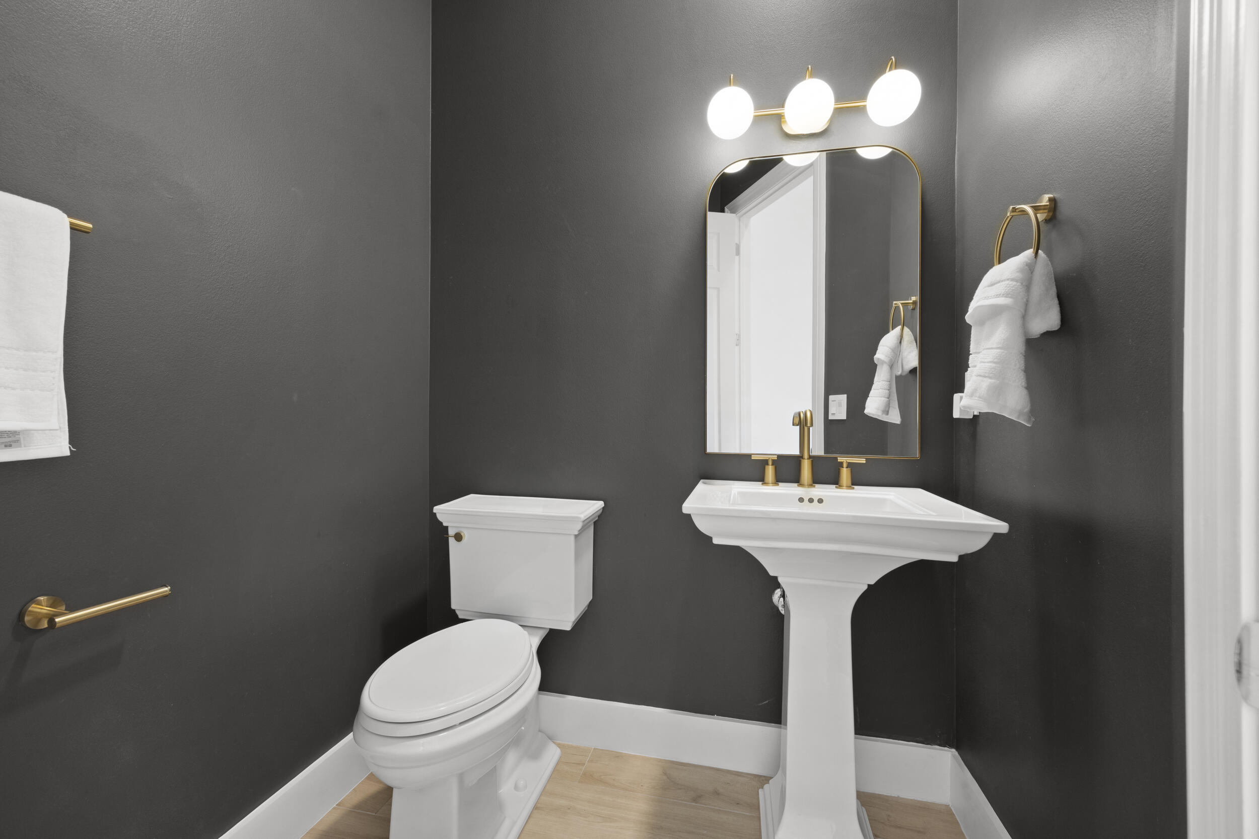 Powder room featuring a white pedestal sink, brass-finish faucet, arched mirror, three-globe light fixture, and wood-finish flooring