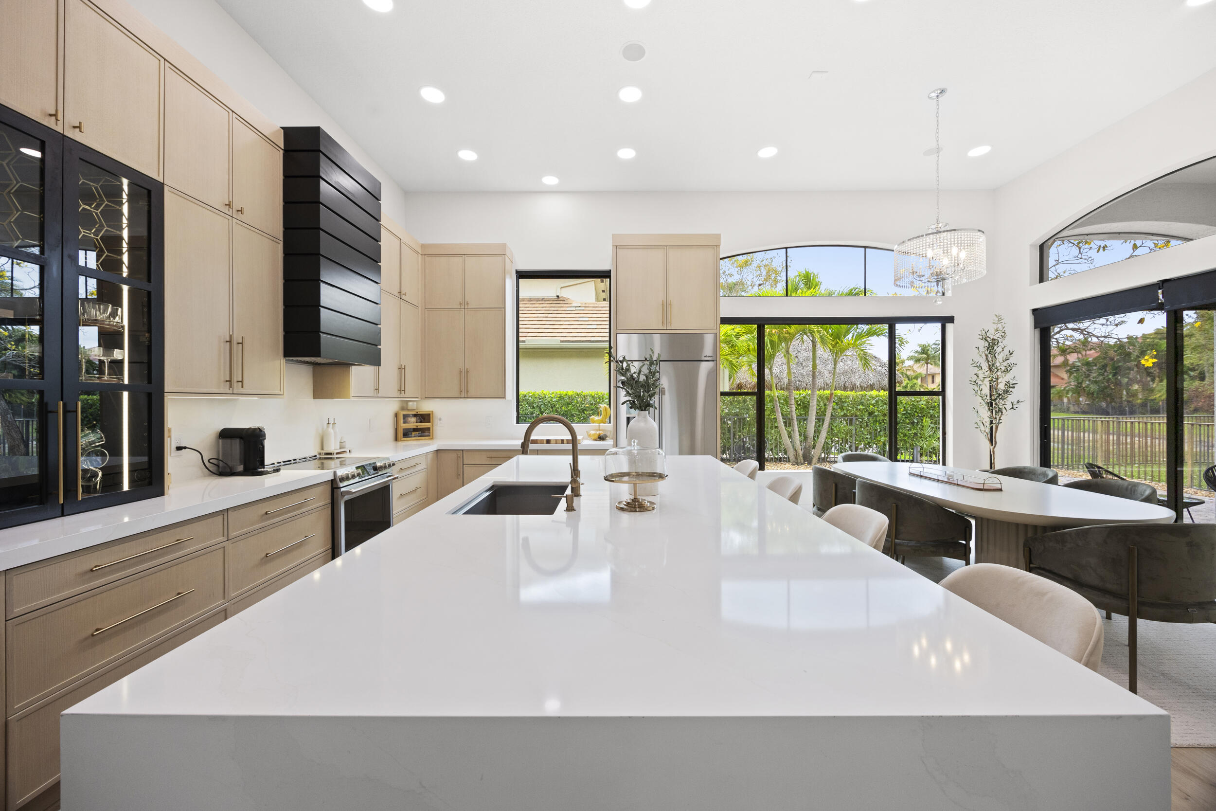Kitchen featuring light wood-finish cabinetry, a large waterfall-edge quartz island, a high-contrast black range hood, glass-front display cabinetry, and oversized arched windows