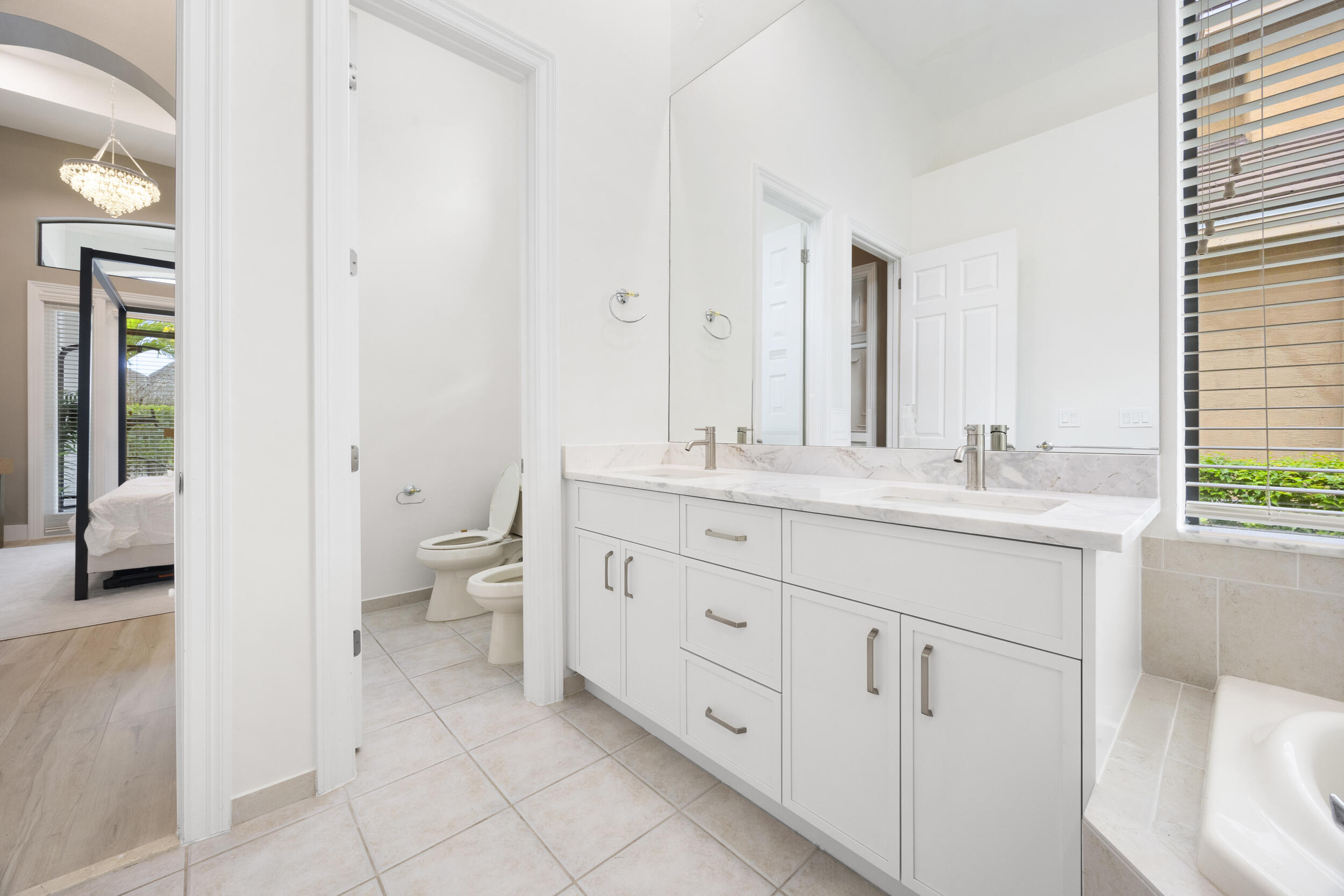 Bathroom featuring a dual-sink vanity with light-toned stone countertops, white cabinetry with brushed-metal hardware, a large wall-mounted mirror, and tile flooring