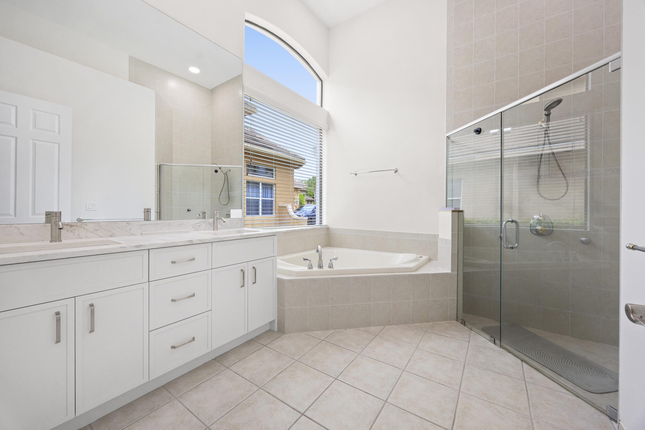 Bathroom featuring a dual-sink vanity with light stone countertops, a large glass-enclosed shower, a deep soaking tub set within an arched window alcove, and tile flooring