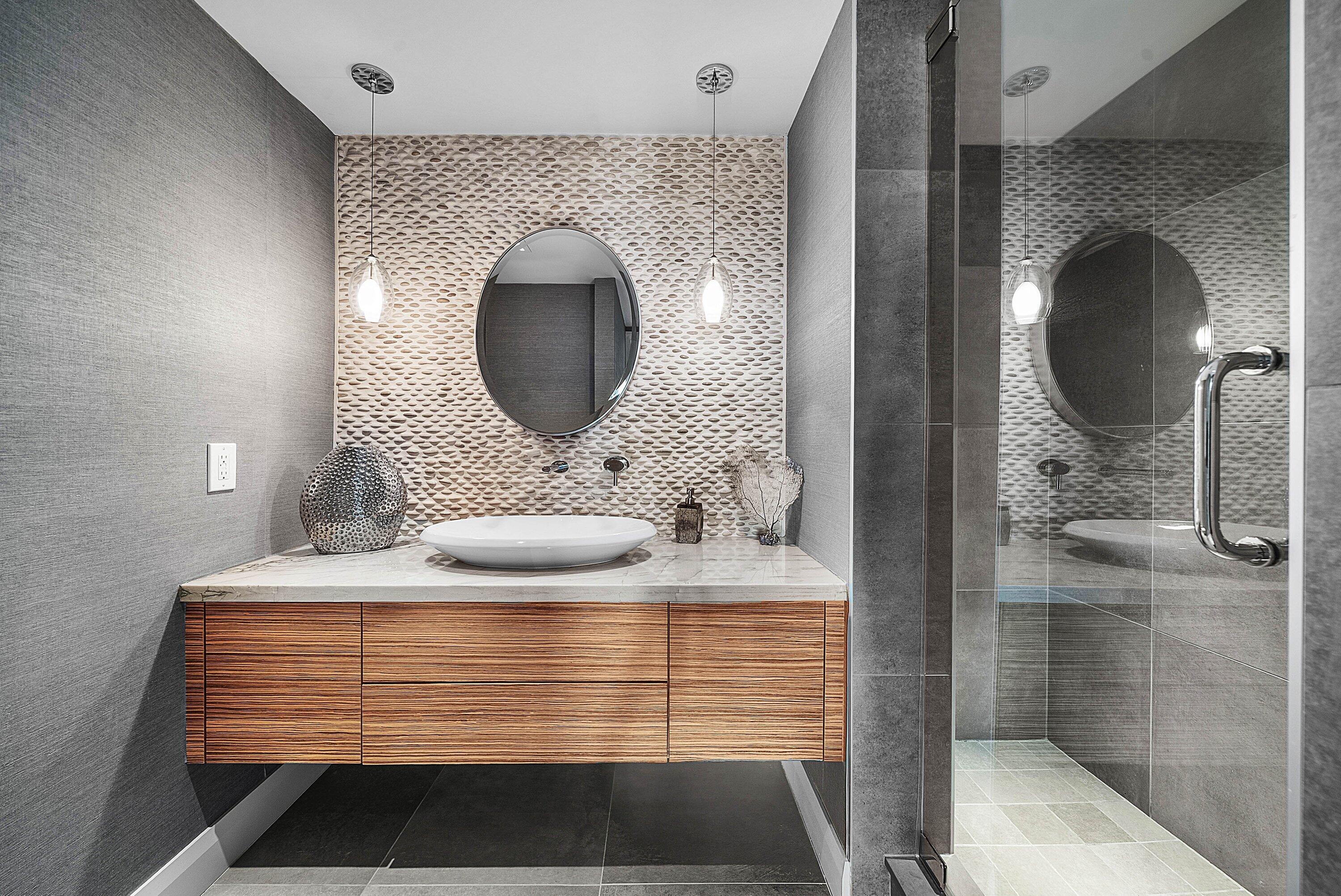 Floating wood-finish vanity with a vessel sink and stone countertop, pebble-texture accent wall, dual pendant lighting, and a glass-enclosed shower with large-format wall tiles